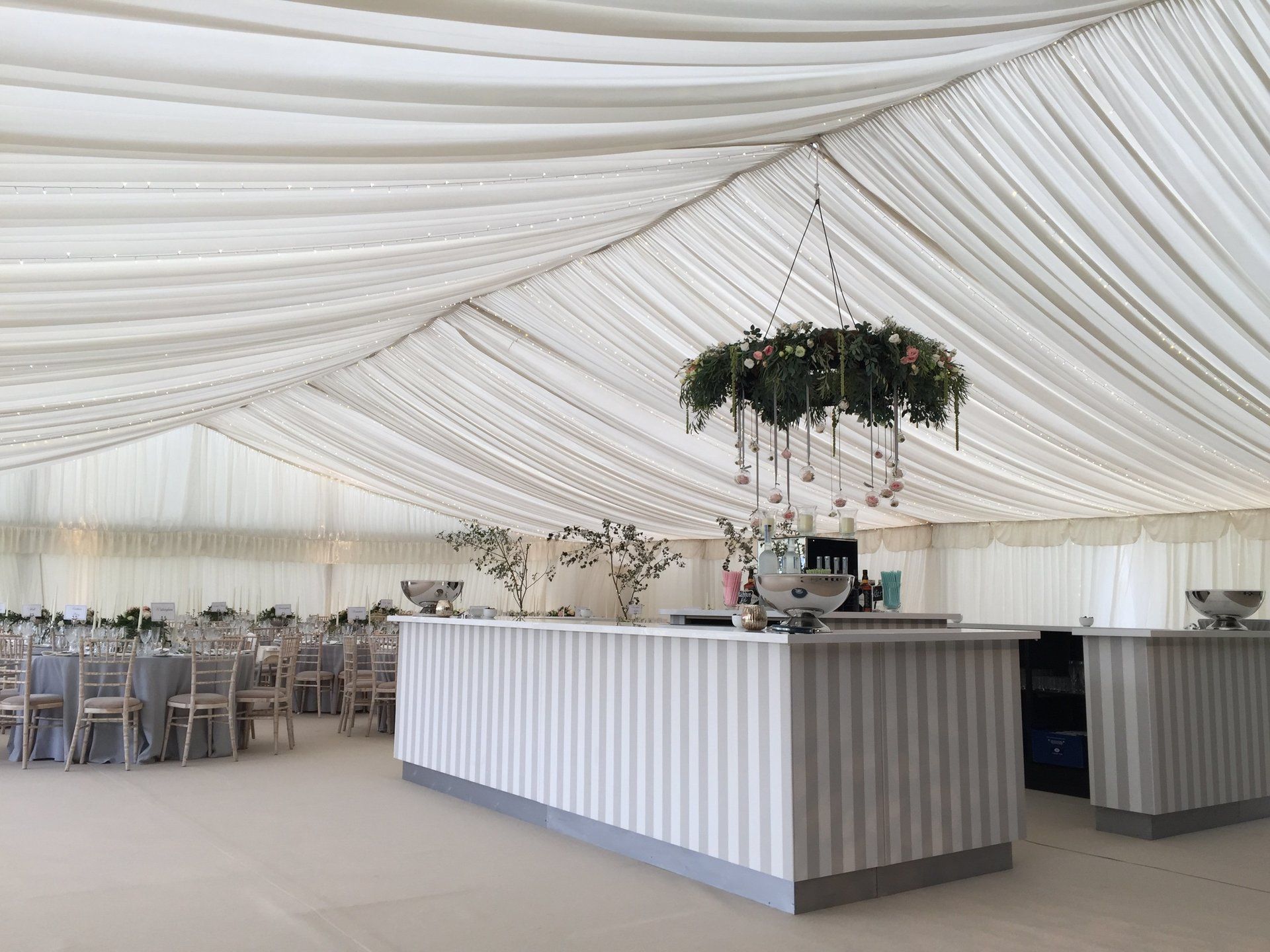 White tent reception with striped bar, hanging floral decor, and round tables set for an event