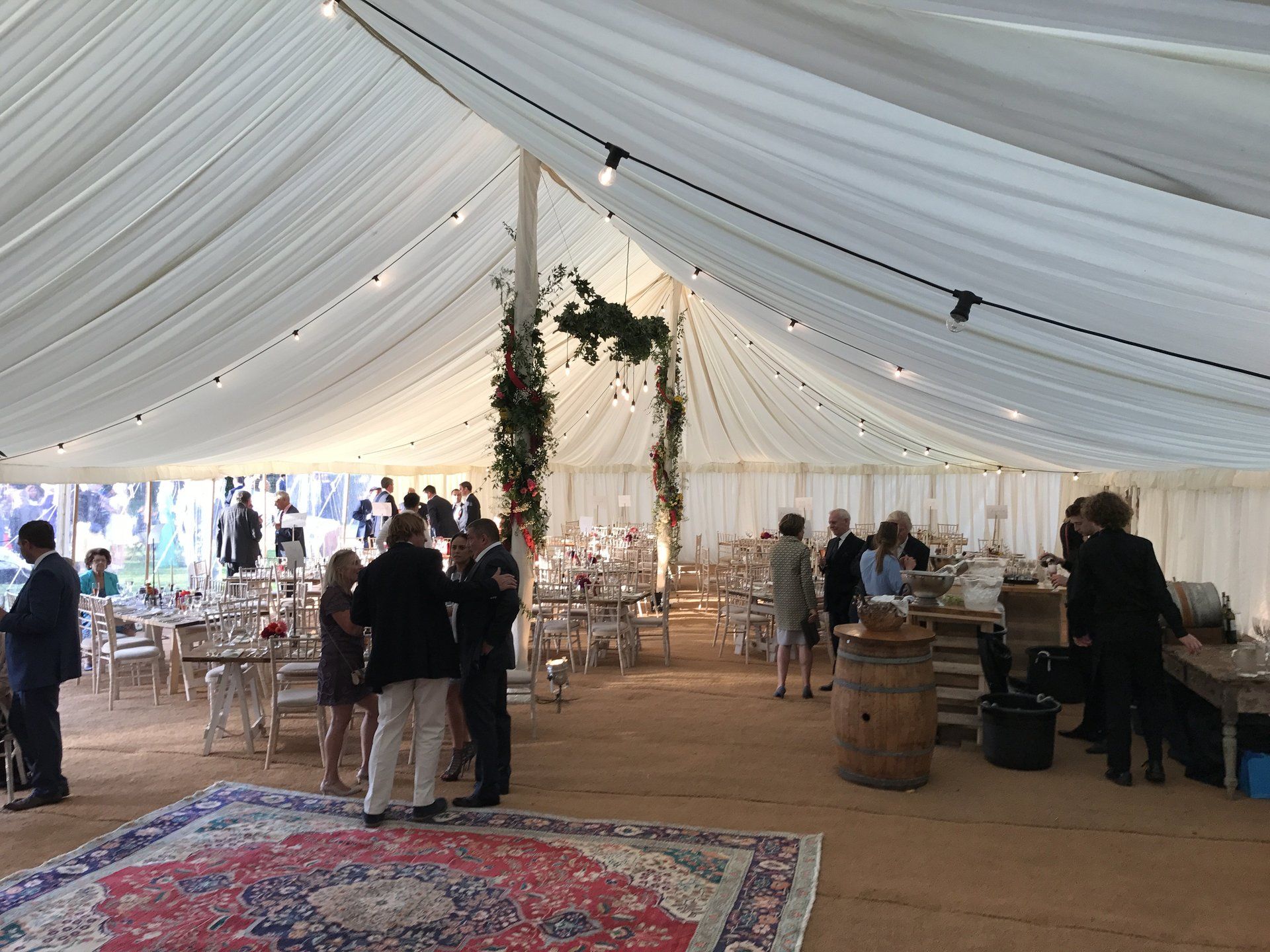 Inside a white event tent with guests, tables, string lights, and a decorated arch on a patterned rug.