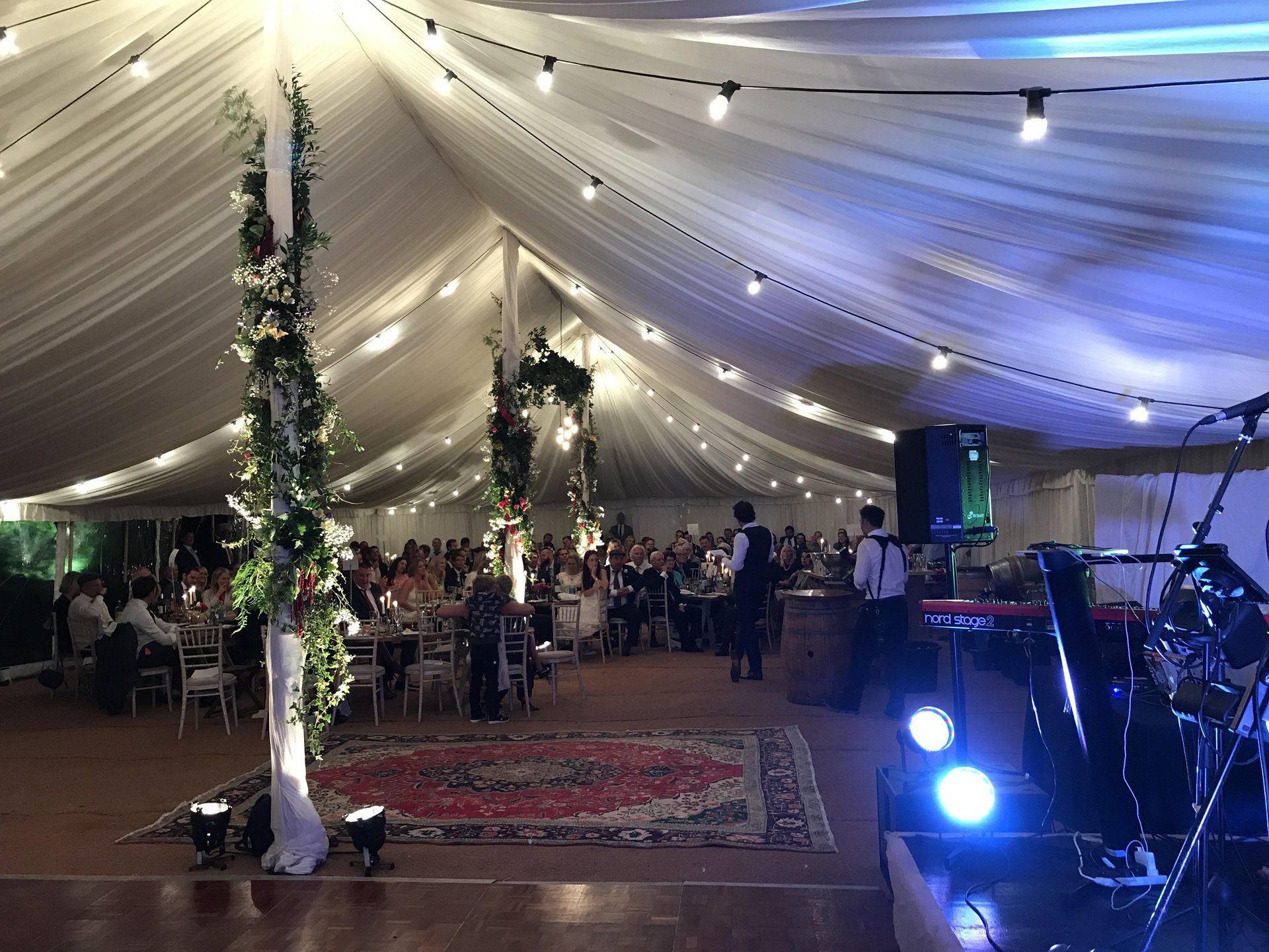 Indoor tented event with guests seated at tables, string lights, and a band stage at the right
