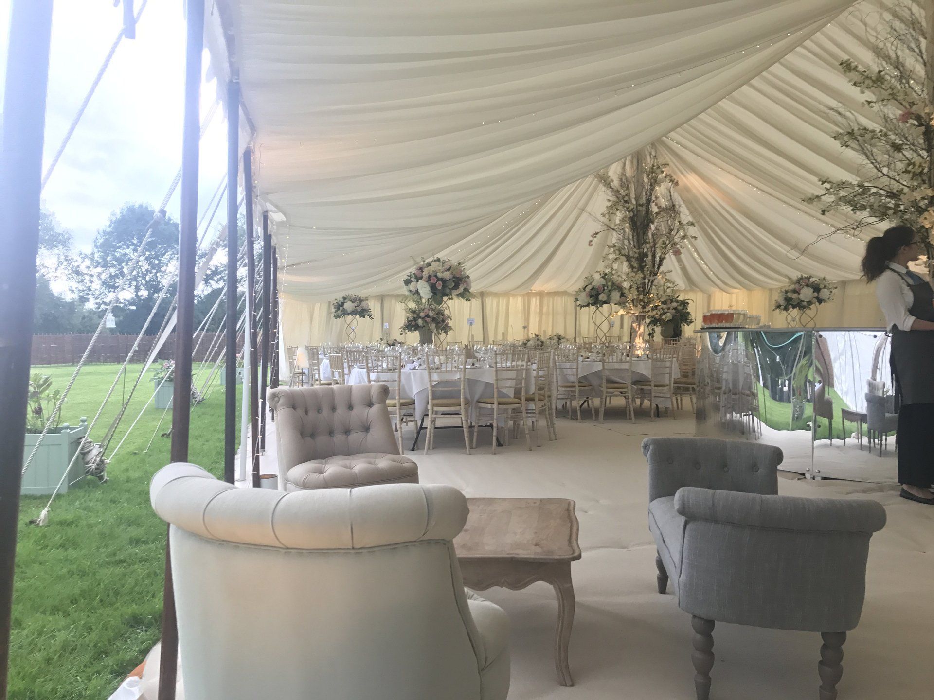 White tented outdoor reception with round tables, beige and gray chairs, and a grassy field beside it