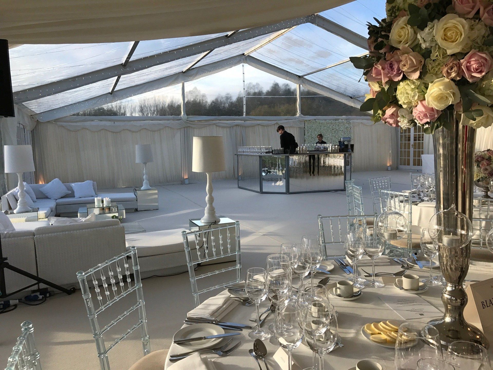 Elegant tented event setup with white chairs, set tables, and a floral centerpiece by large windows