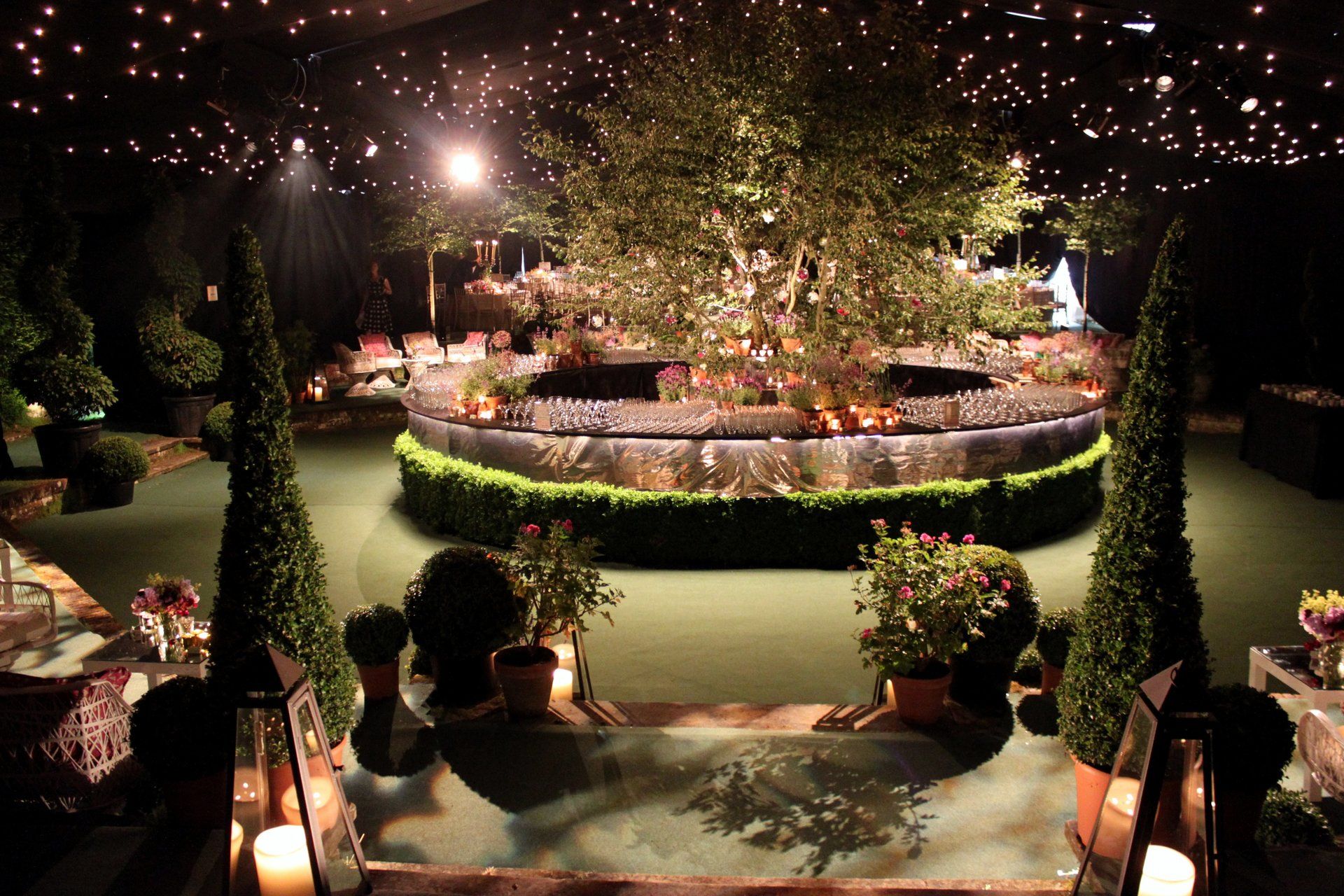 Lit garden courtyard with a central fountain, trimmed hedges, and warm evening lights under trees