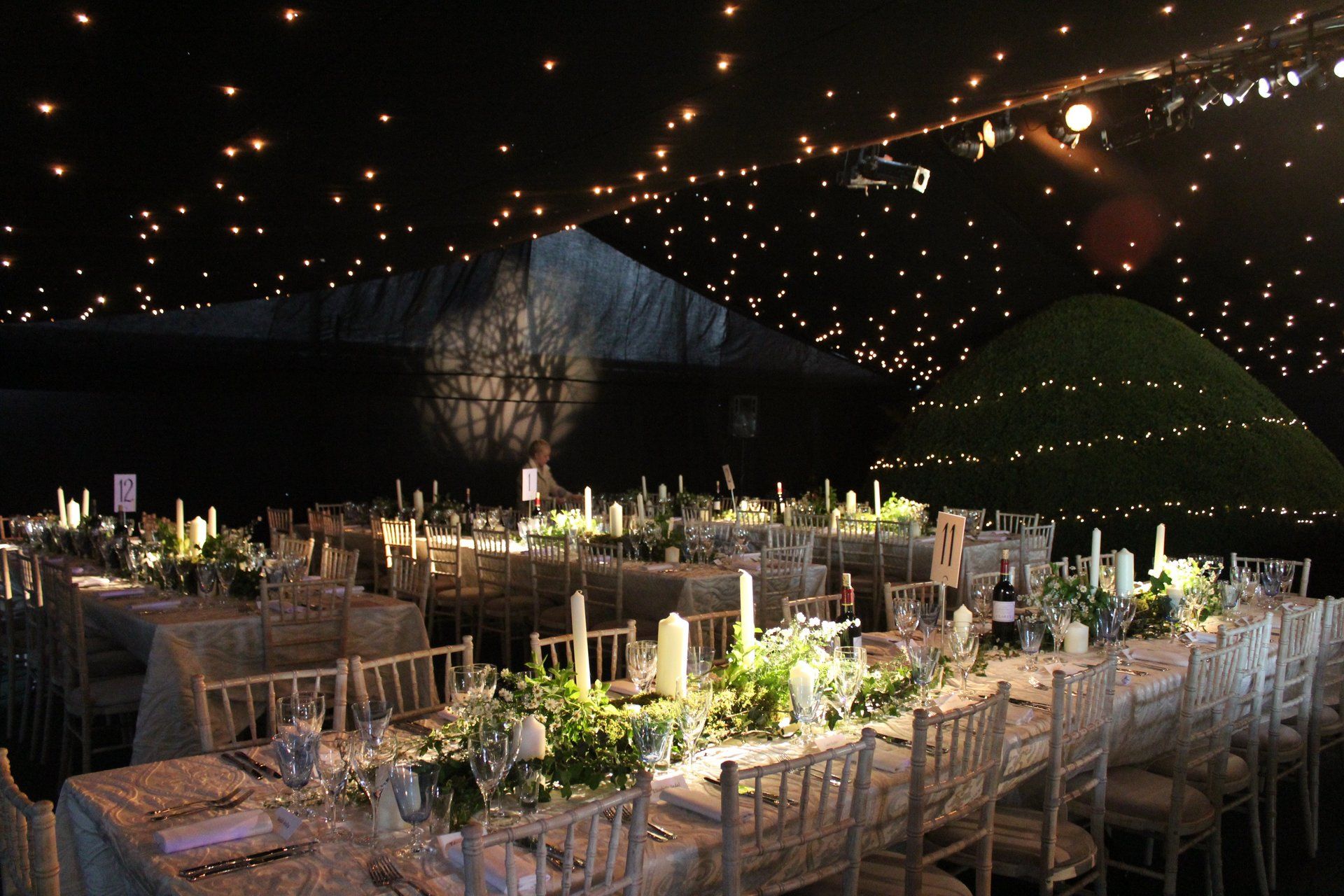 Elegant outdoor banquet with long tables, white chairs, and string lights under a dark tent-like canopy at night