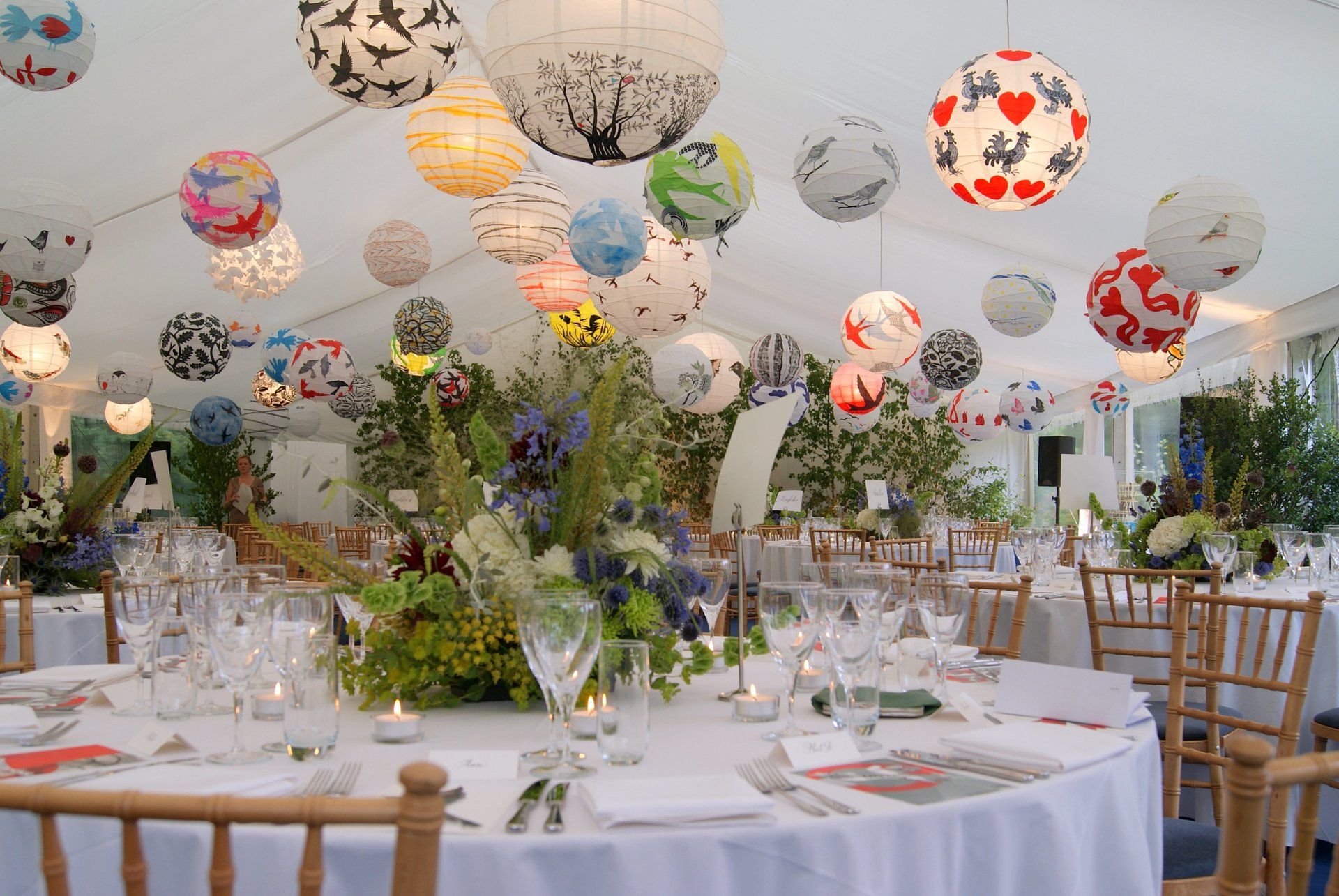 Decorated banquet hall with round tables, white chairs, floral centerpieces, and hanging paper lanterns under a tented ceiling