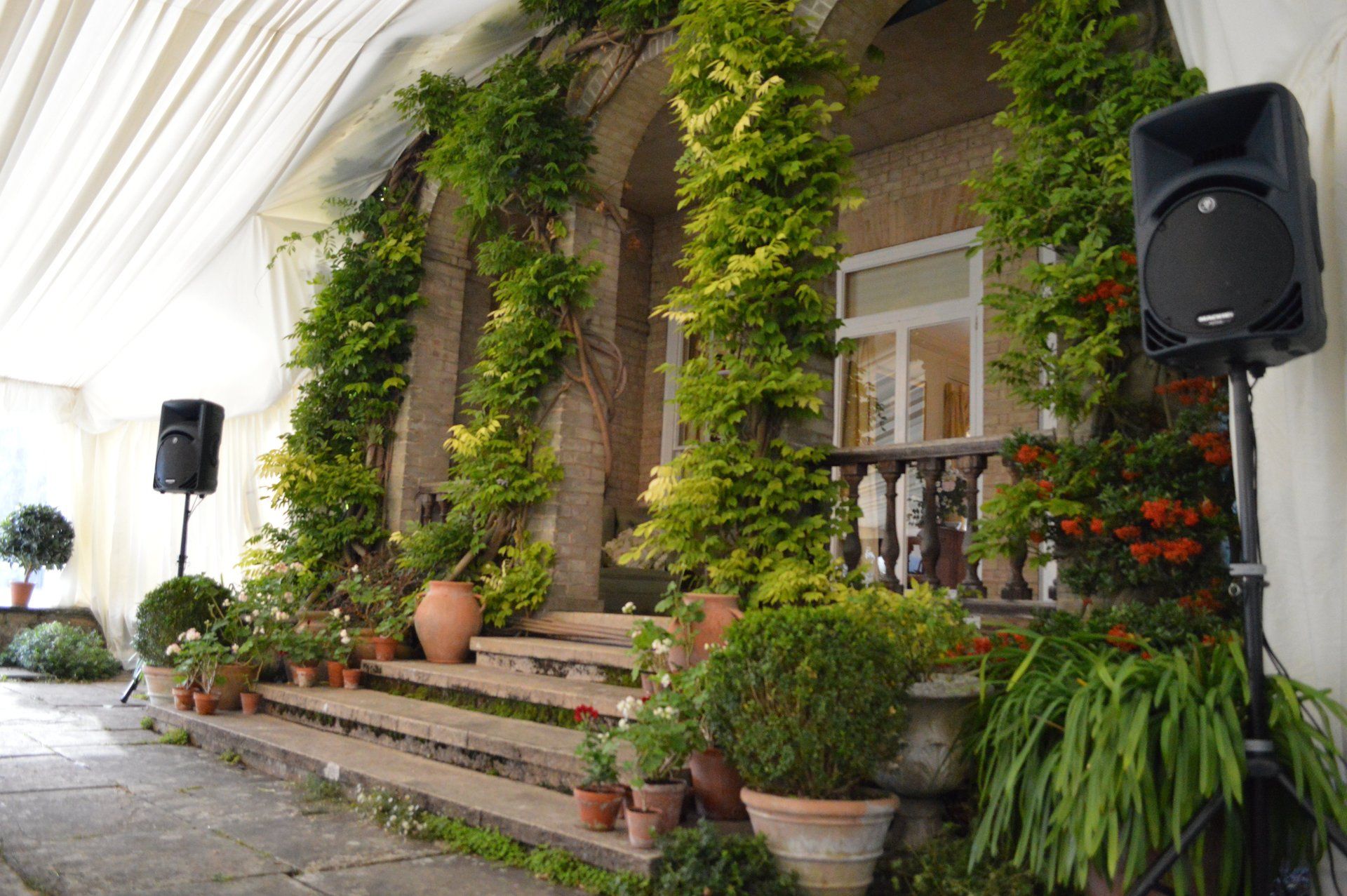 Flower-covered building entrance with potted plants and large black speakers under a white canopy