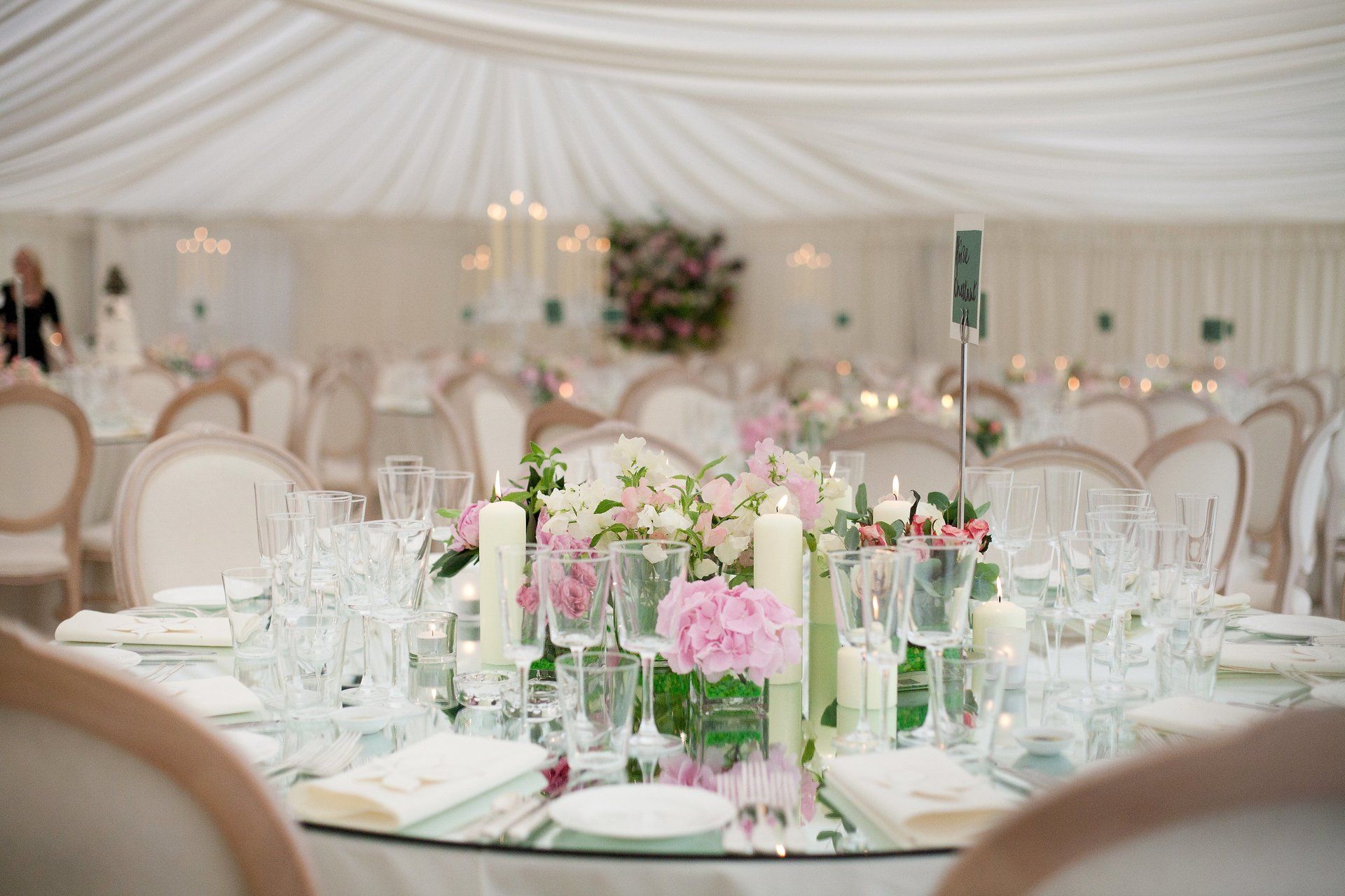 Elegant wedding reception table with pink floral centerpiece, candles, and glassware under a white draped tent