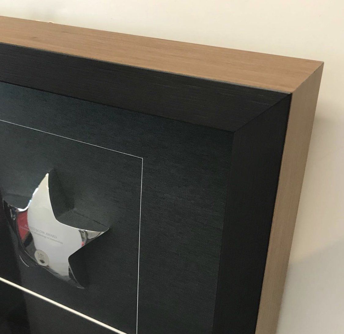 A framed artwork with a dark, textured backing featuring a silver star element, encased in a wood and black border.