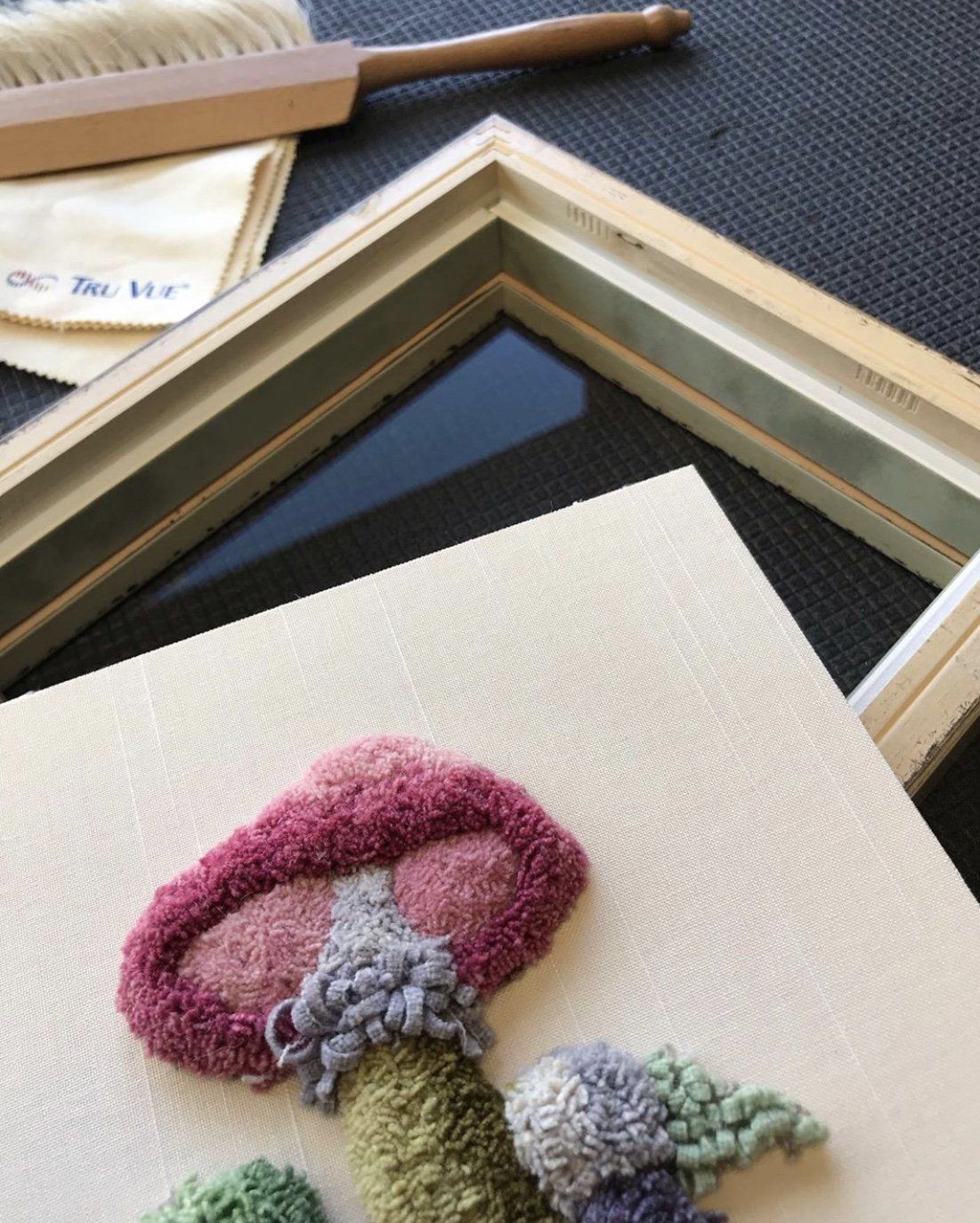 A textured punch needle mushroom art piece sits beside an empty wooden picture frame on a dark surface.