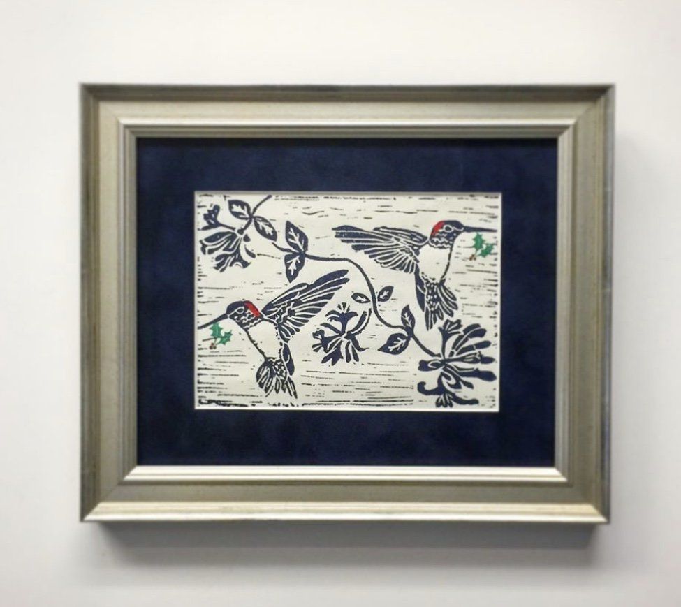 A framed linocut print shows two hummingbirds with red heads hovering near floral vines against a cream background.