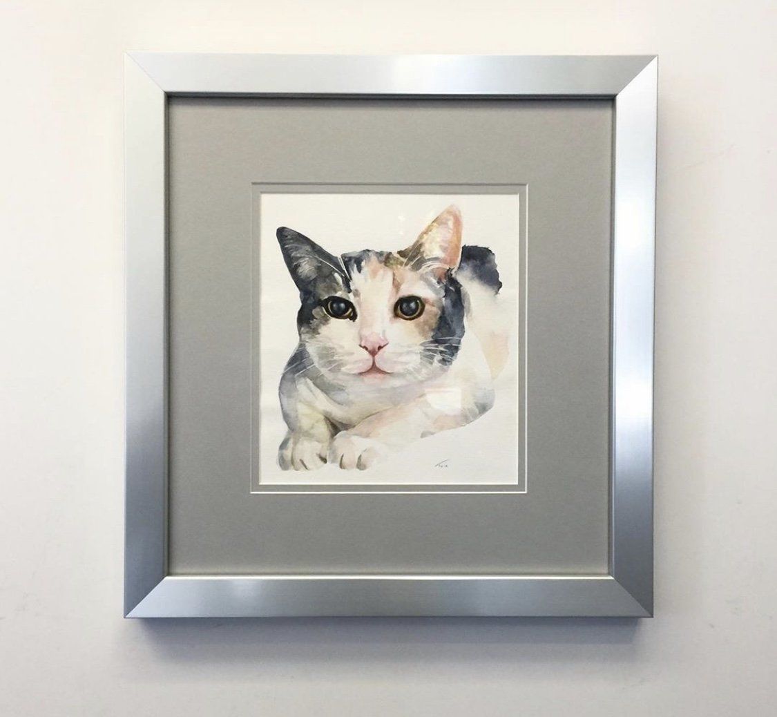 A framed watercolor painting of a calico cat with large eyes and folded paws, hanging on a plain white wall.