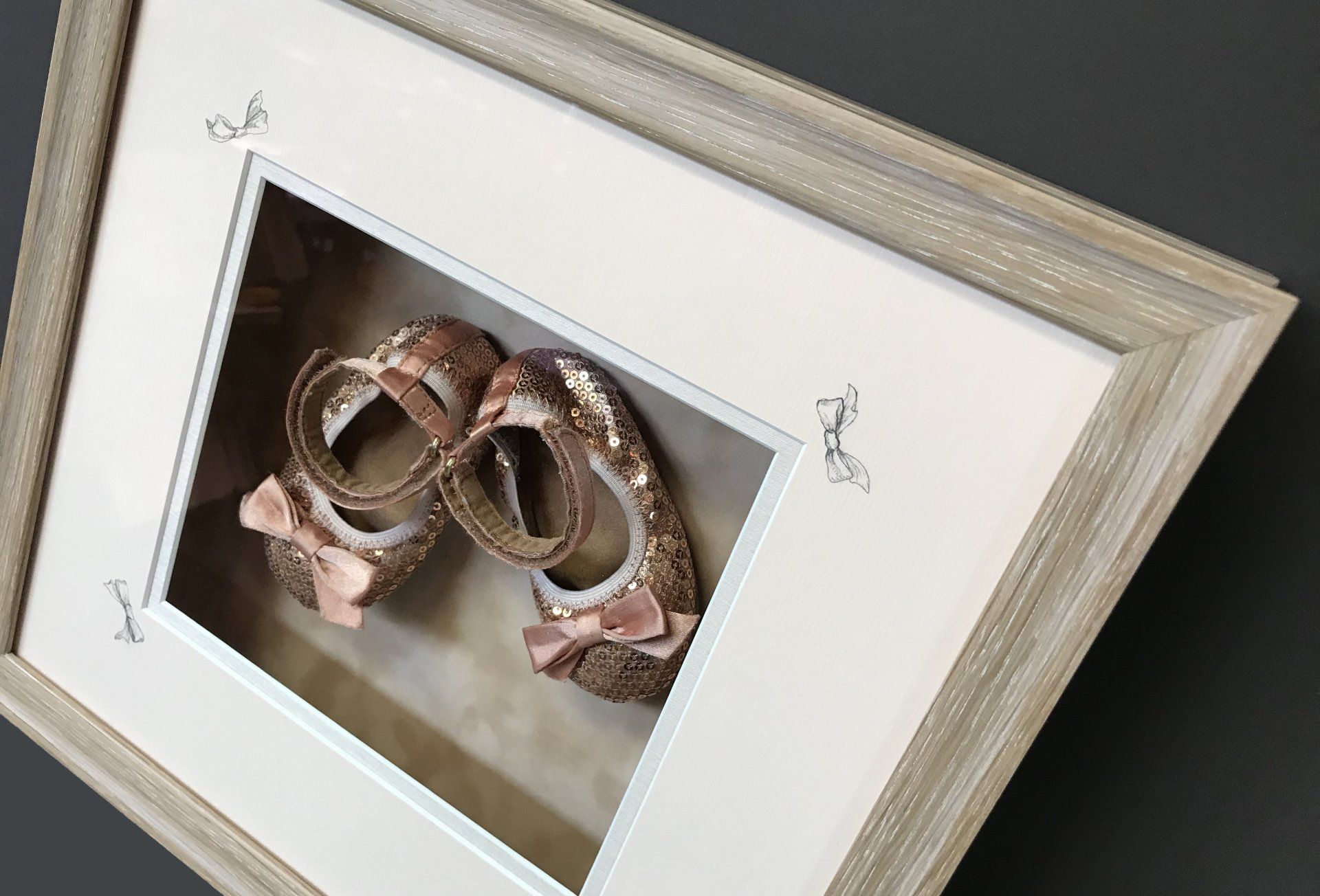 A framed shadow box containing a pair of small, sparkly rose gold baby shoes with bows.