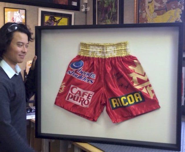 A person holding a large framed display of red, gold-trimmed boxing trunks featuring various sponsor logos and a signature.