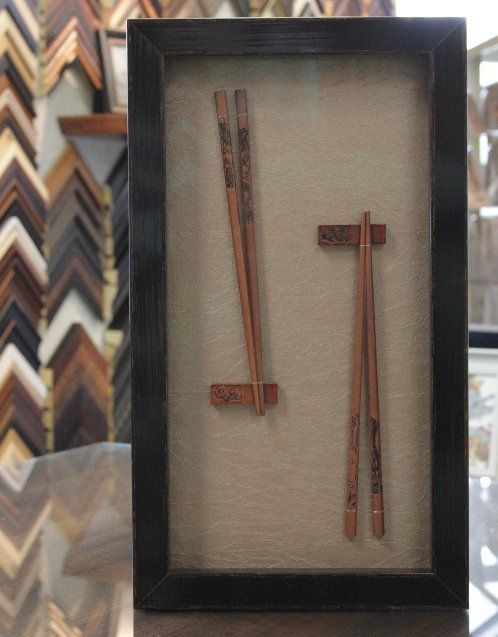 A shadow box display featuring two sets of wooden chopsticks, each resting on a small, matching holder, against a tan mat.