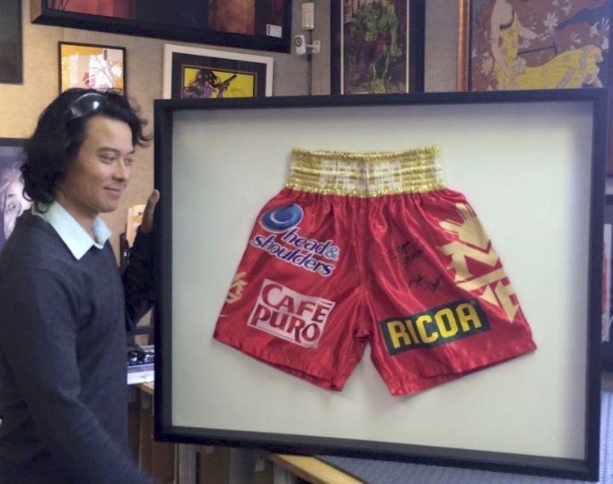A person holding a large shadow box frame containing a pair of red boxing trunks with various logos, displayed indoors.