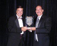 Two men in tuxedos holding an award, smiling at the camera, on a stage with a dark curtain backdrop.