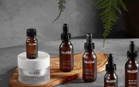 Five Medilis skincare bottles on a wooden tray and white pedestal against a gray wall with greenery