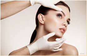 Woman with slicked-back hair posing as gloved hands frame her face in a beauty treatment setting