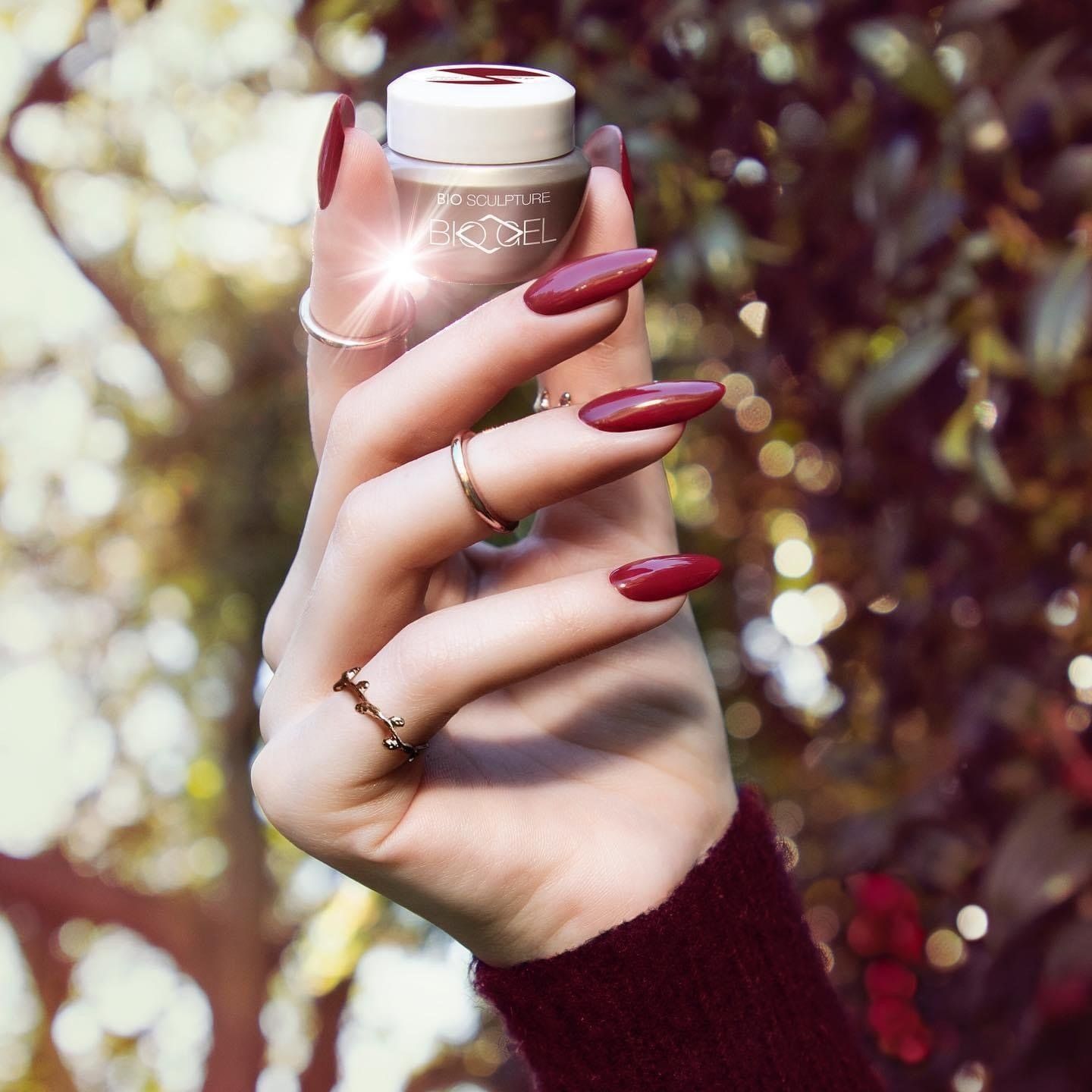 Hand holding a beige cosmetic bottle with long burgundy nails against a blurred leafy background