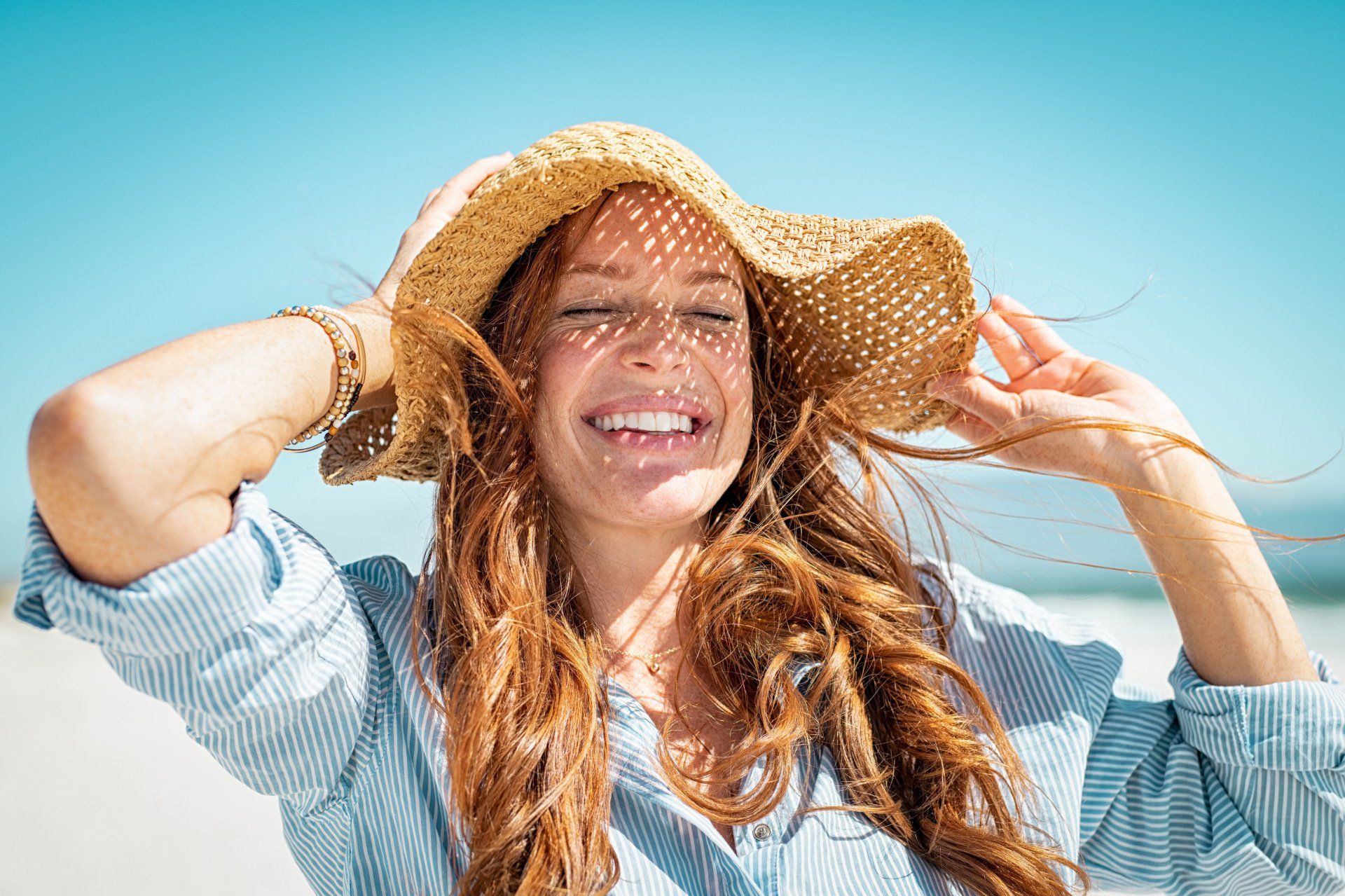 Smiling woman in a straw hat adjusting it on a sunny beach with blue sky backdrop