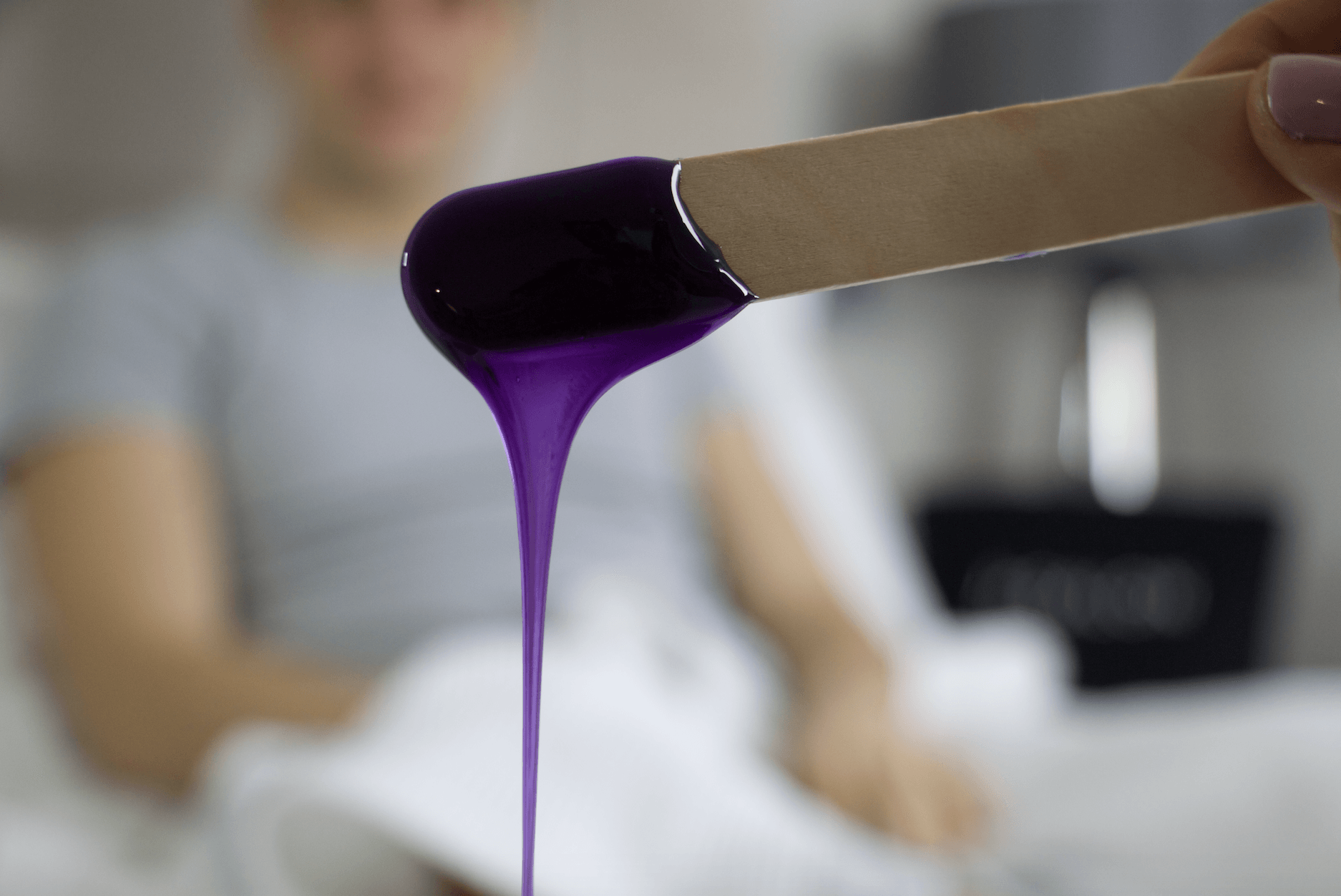Purple wax dripping from a wooden spatula over a blurred spa treatment table