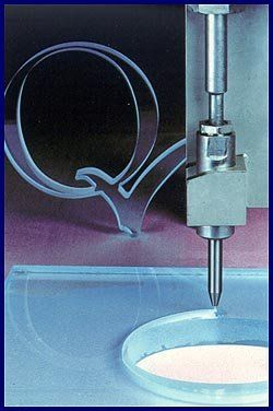 Metal nozzle dispensing material into a blue circular mold on an industrial machine, with a looped tube beside it