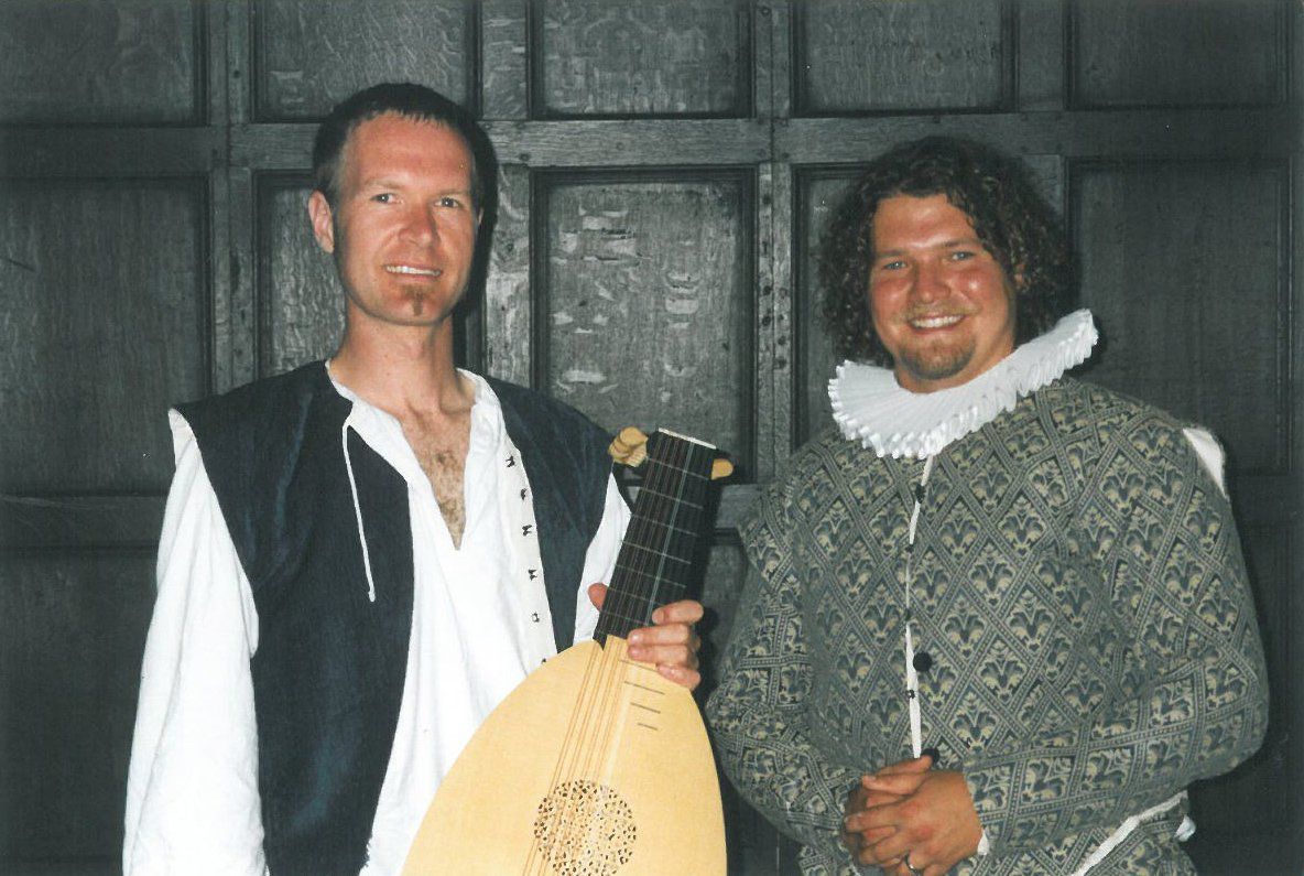 Two men standing next to each other one holding a guitar and the other holding a lute