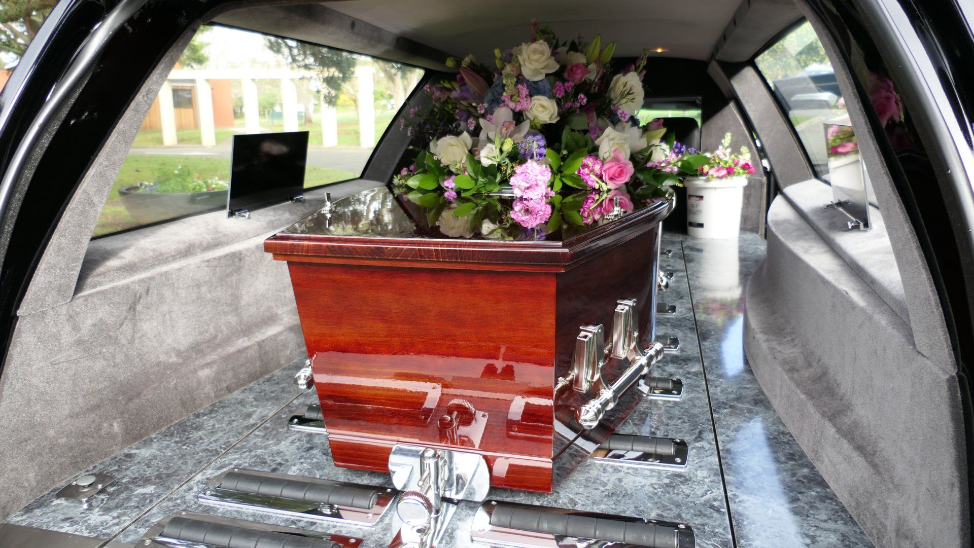 A flower-adorned wooden casket inside a hearse with the rear door open.