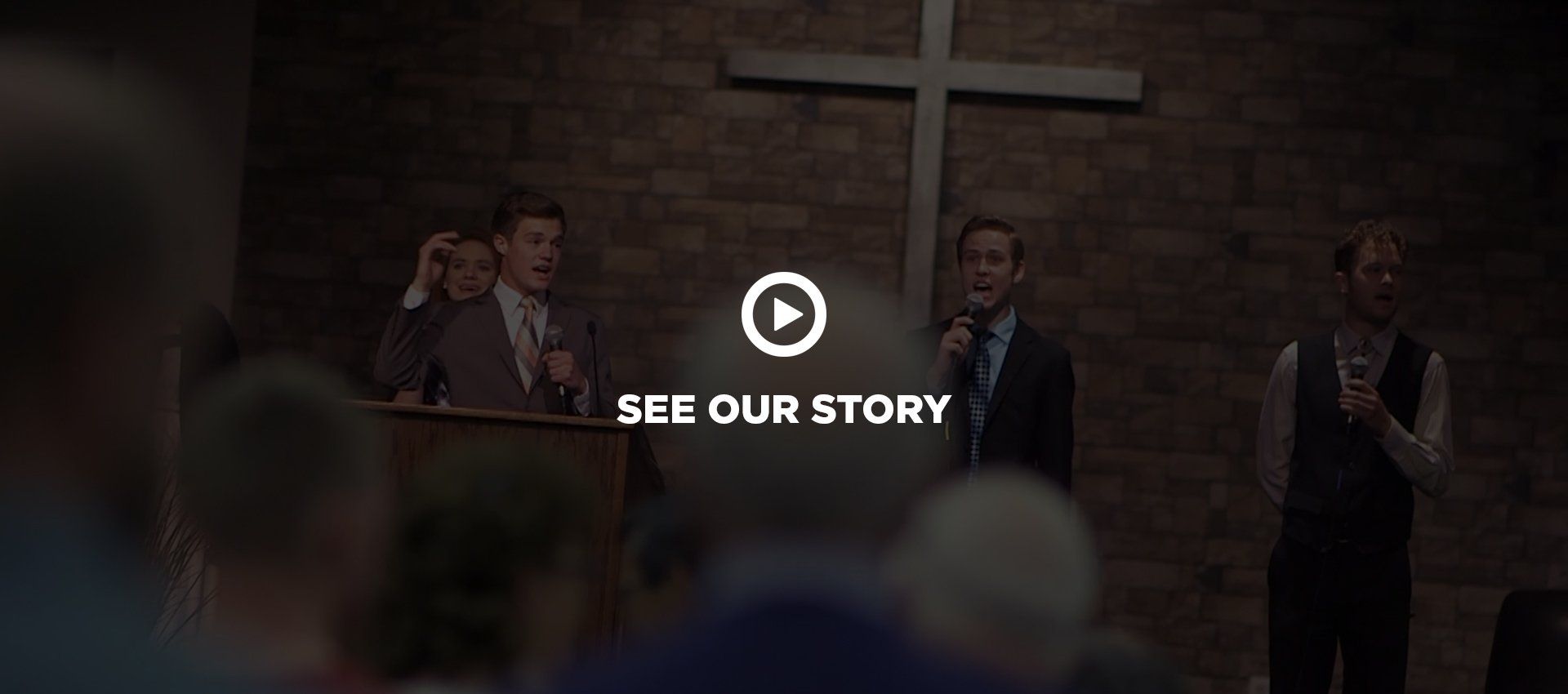 Two young gentlemen on a stage speaking with a cross behind them on the wall.
