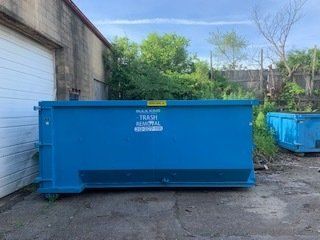 A bright blue dumpster sits on a paved lot beside a building with white garage doors and overgrown trees in the back.