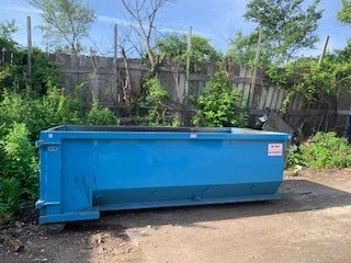 A bright blue roll-off dumpster sitting on a dirt lot in front of a wooden fence and overgrown vegetation.