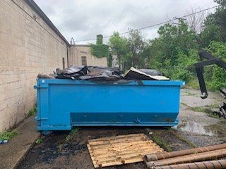 A large, blue metal dumpster filled with construction debris sits next to a beige building wall on a gravel lot.