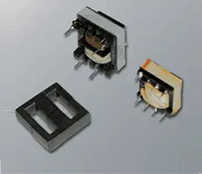 Tre trasformatori elettrici di diverse dimensioni e un nucleo nero su una superficie grigia.