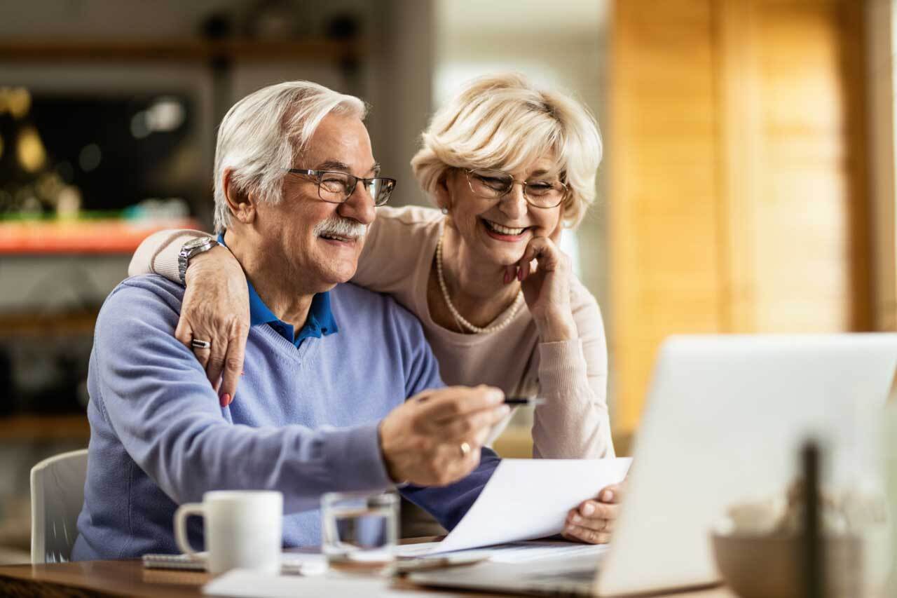 An elderly couple is sitting at a table looking at a laptop computer.