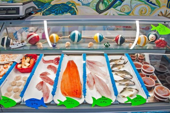 Seafood display case: salmon, fish fillets, whole fish, and prepared foods with decorative buoys above.