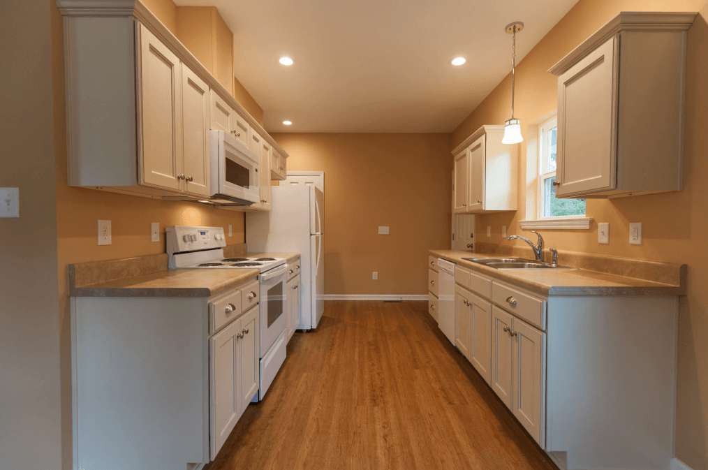 Empty galley kitchen with white cabinets, tan walls, and wood floors viewed from the doorway