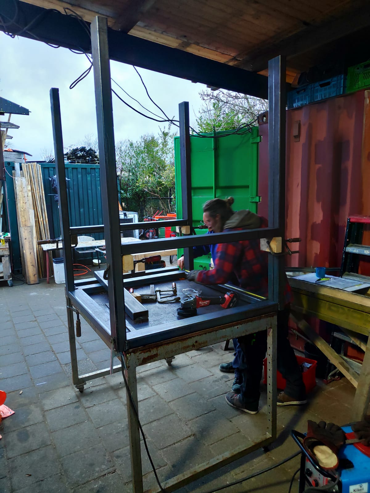 Een persoon last buiten, onder een afdak, het metalen frame van een werkbank.