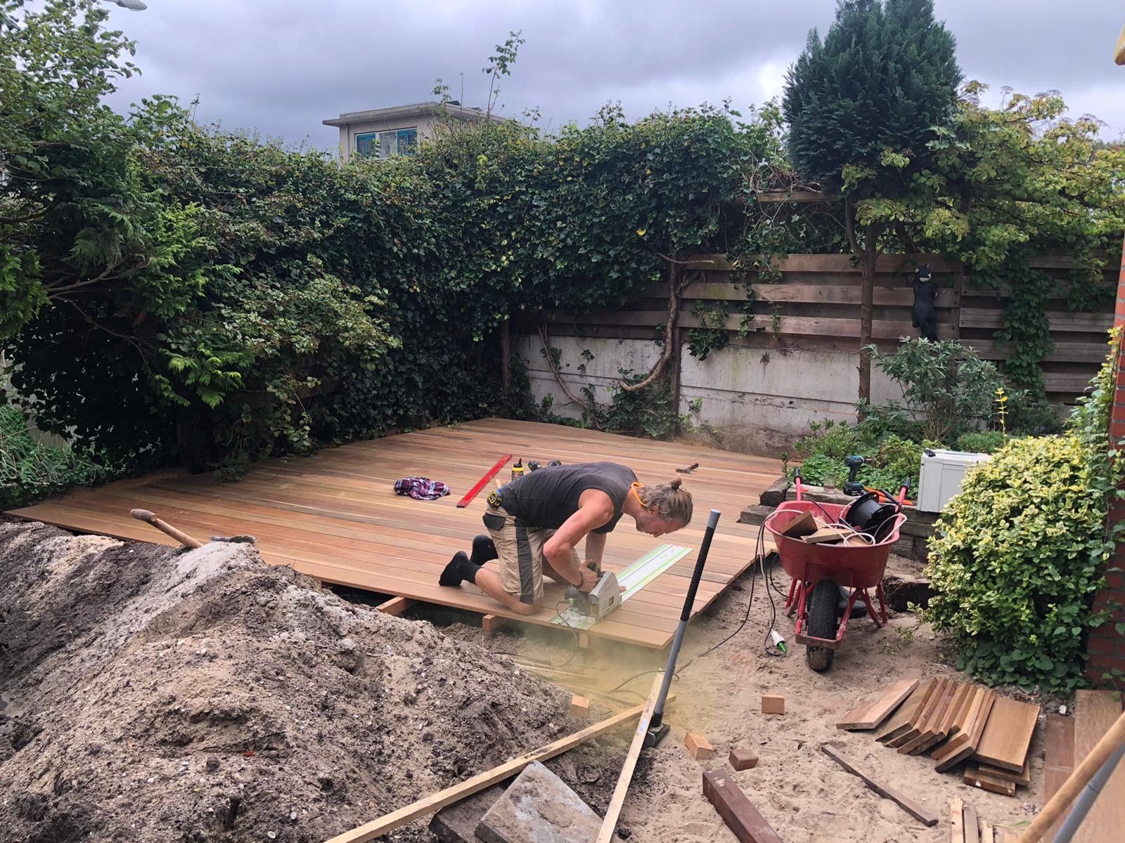 Een persoon bouwt een houten terras in de achtertuin, met gereedschap en materialen zichtbaar.
