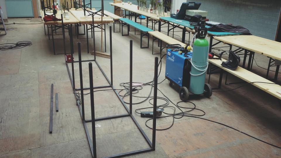 Een metalen frame wordt buiten gelast, met tafels en lasapparatuur in de buurt.