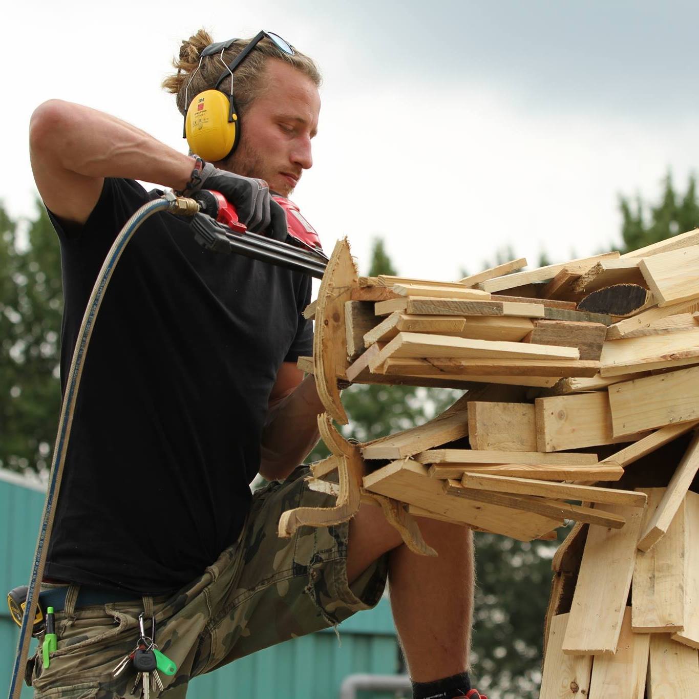 Een man gebruikt gereedschap om buiten aan een houten sculptuur te werken, met gehoorbescherming en handschoenen aan.