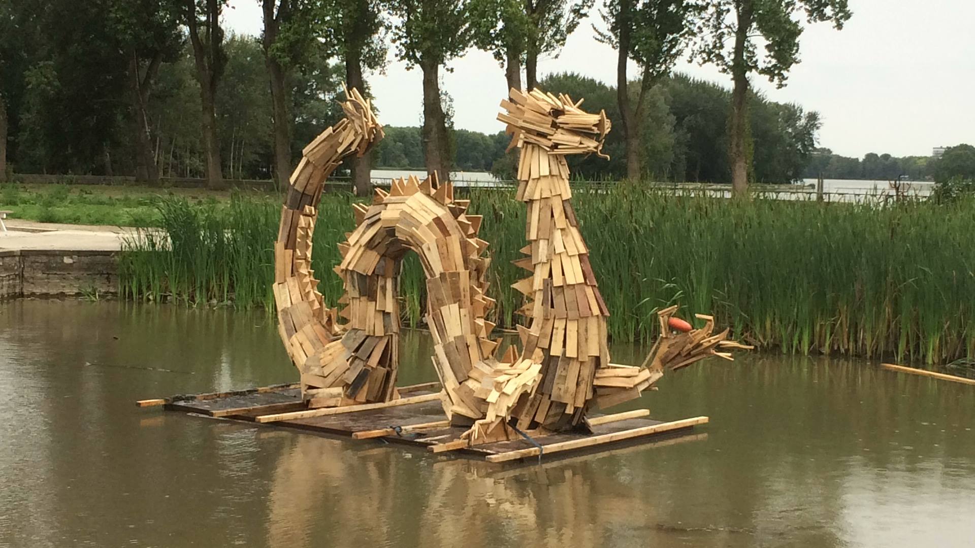 Een houten drakensculptuur drijft in het water, met riet en bomen op de achtergrond.