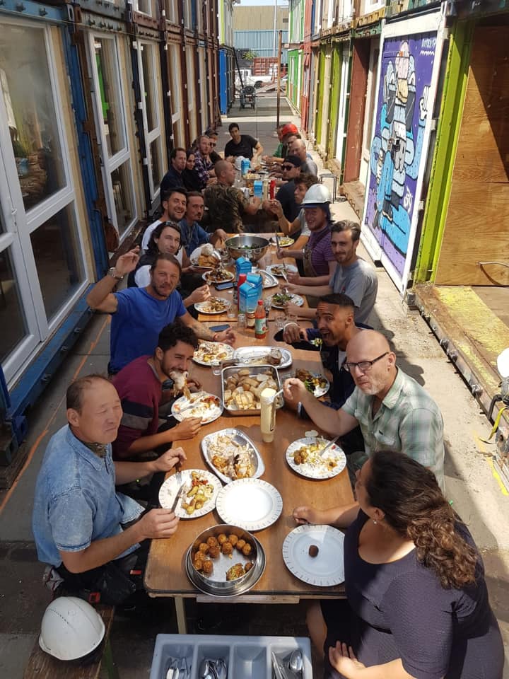 Een lange tafel waaraan mensen buiten eten. Aan weerszijden staan gebouwen. die eten en lachen.