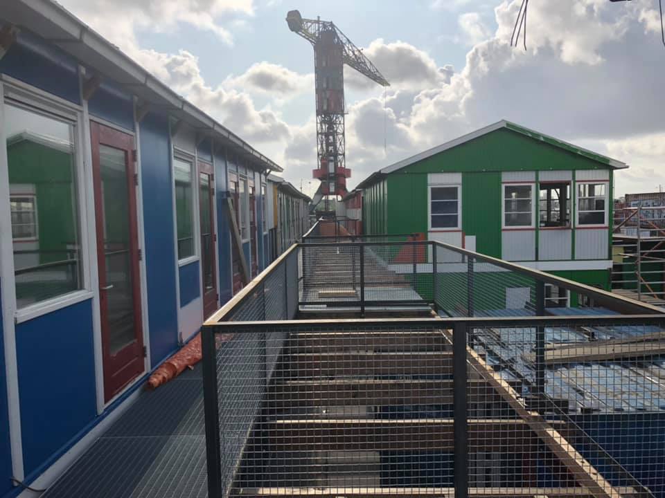 Metalen loopbrug tussen kleurrijke modulaire gebouwen op een bouwterrein; kraan op de achtergrond.
