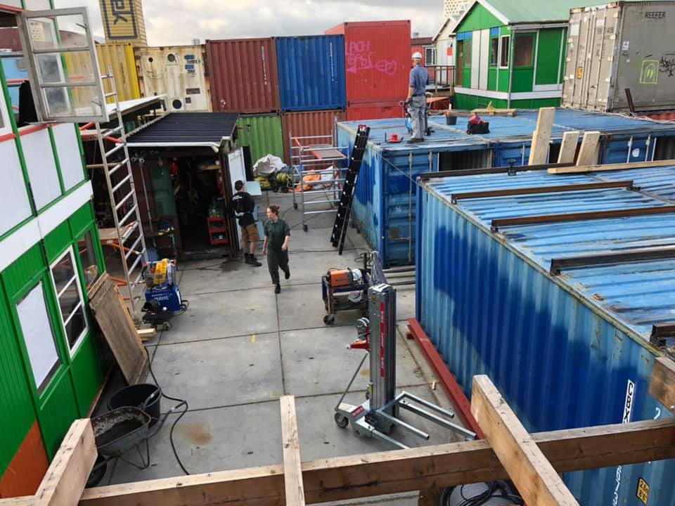 Bouwterrein met zeecontainers in verschillende kleuren; er zijn arbeiders aanwezig.