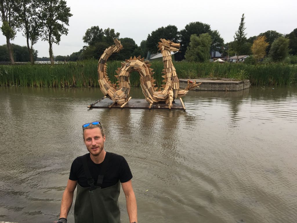 Een man staat in het water, vlakbij een groot houten drakenbeeld dat op een platform drijft.