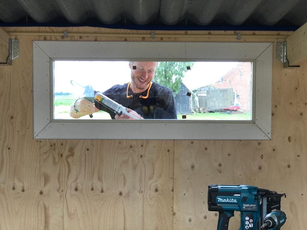 Een man brengt kit aan op een raam in een houten constructie; een spijkerpistool is zichtbaar.