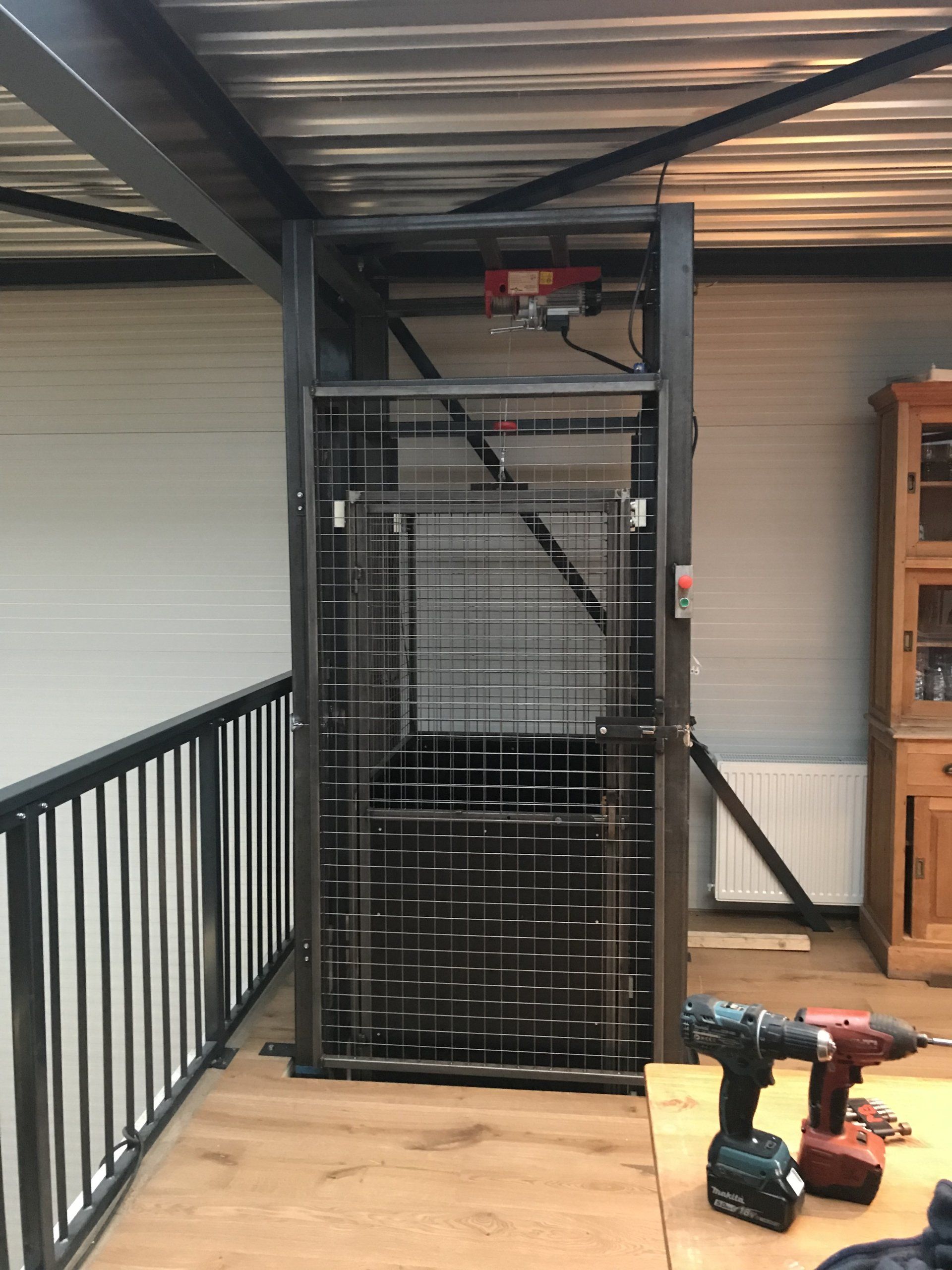 Een platformlift met metalen kooi, geïnstalleerd in een gebouw met een balkon en gereedschap op een tafel.