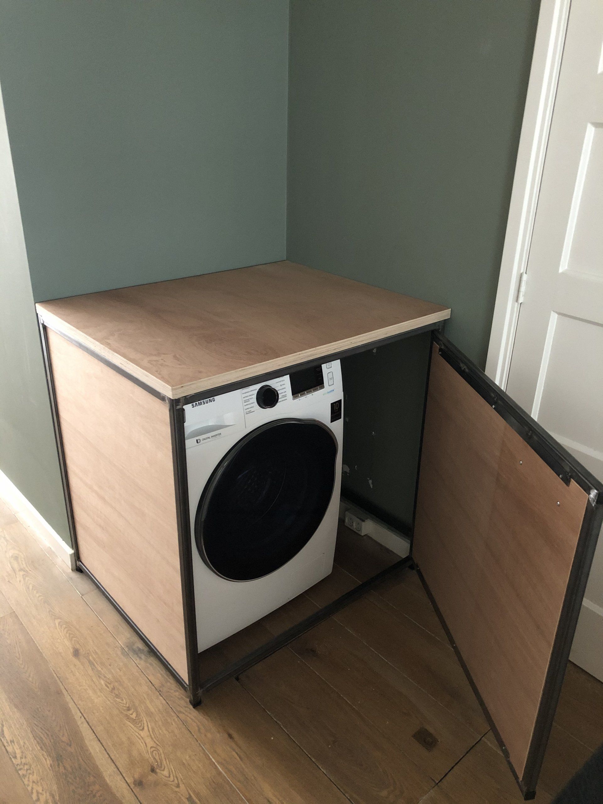Een wasmachine staat in een houten kast met een open deur in een kamer met groene muren en een houten vloer.