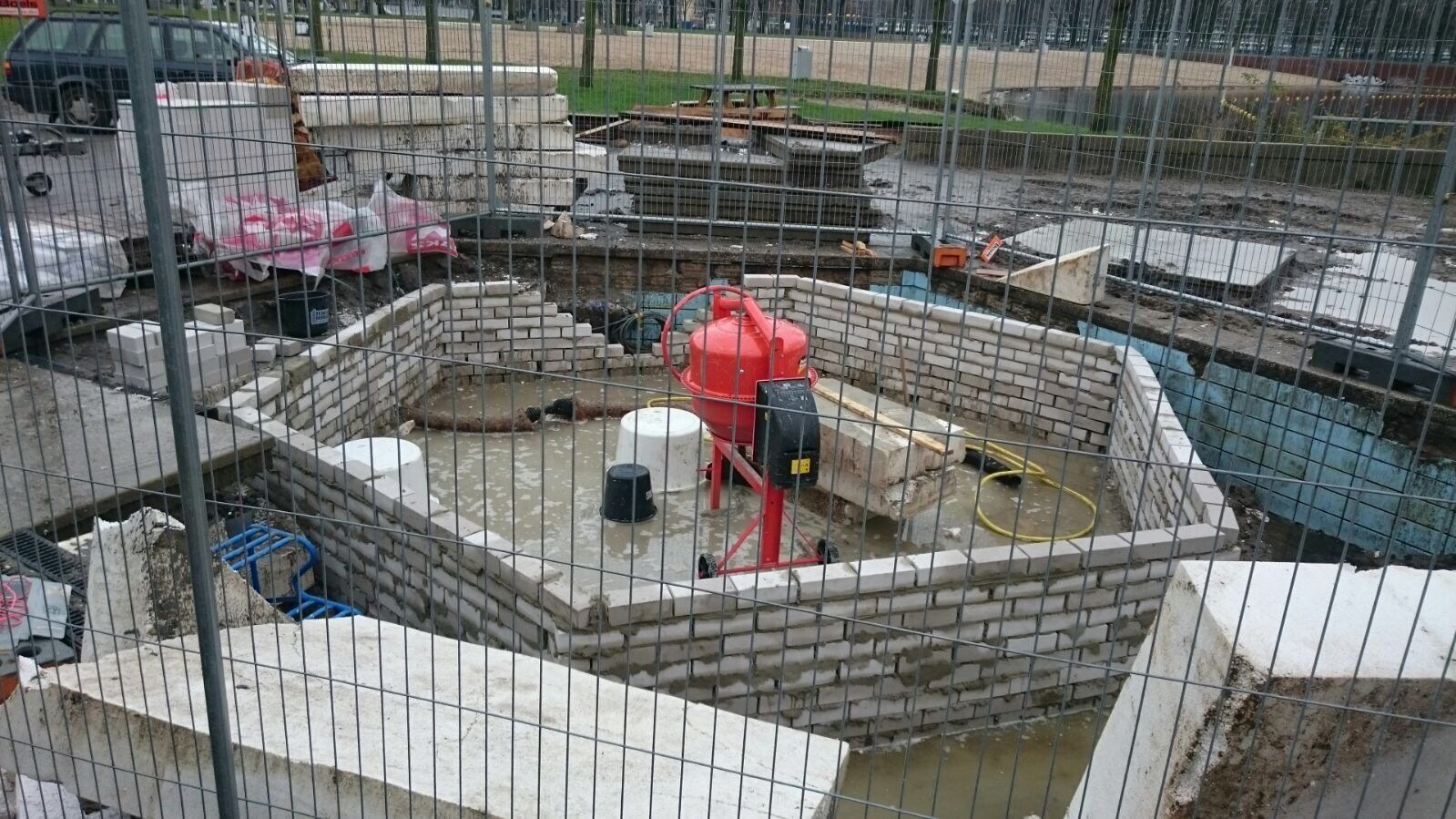 Bouwplaats met een gedeeltelijk gebouwde bakstenen constructie, een rode cementmixer en bouwmaterialen.