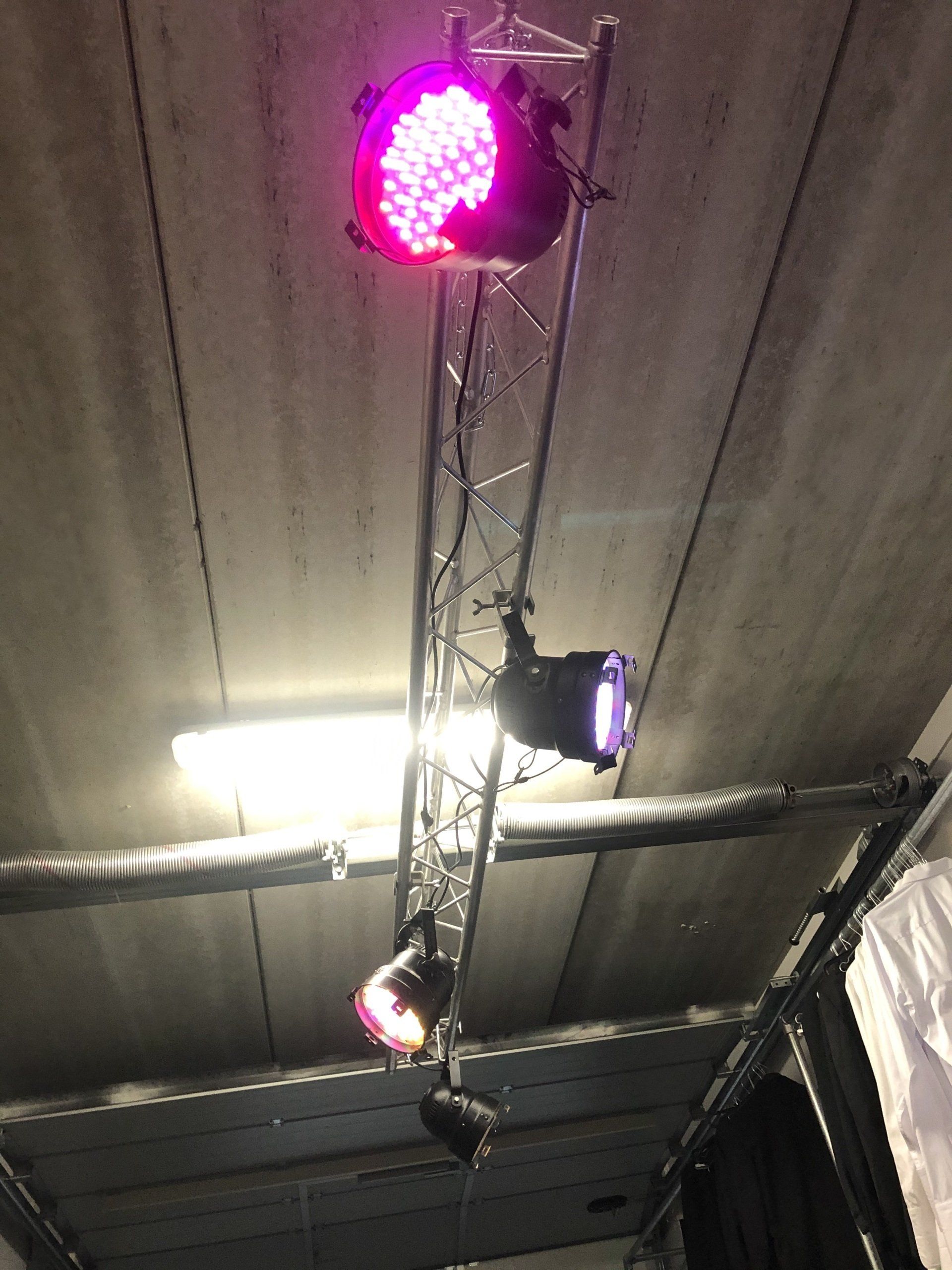 Metalen vakwerkconstructie met podiumverlichting tegen een betonnen plafond; de lampen zijn roze, paars en rood.