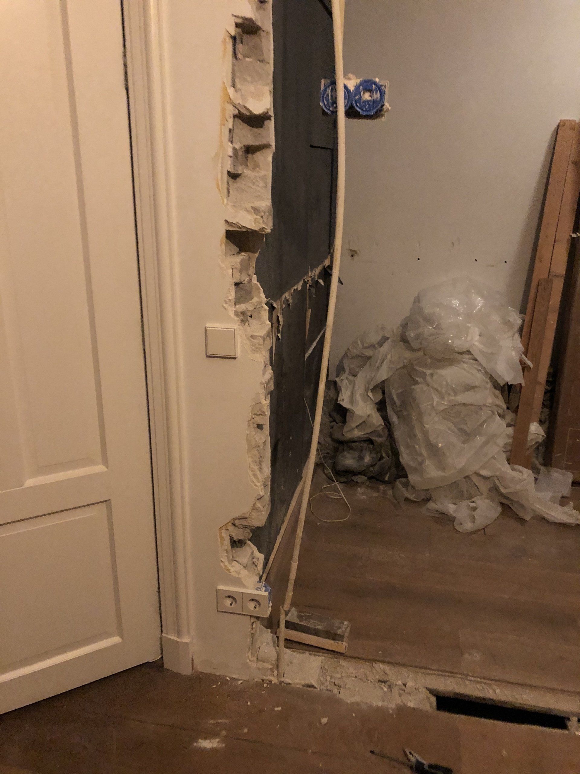 Beschadigde deuropening, muur gesloopt waardoor het interieur zichtbaar is; witte deur links, puin op de vloer.